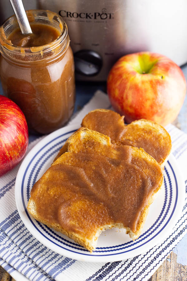 Two pieces of toast slathered in apple butter on a white and blue plate with apples, a jar or apple butter, and a stainless steel Crock Pot in the background.