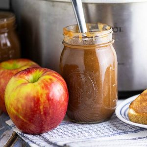 A jar of apple butter next to honeycrisp apples in front of a stainless steel Crock Pot