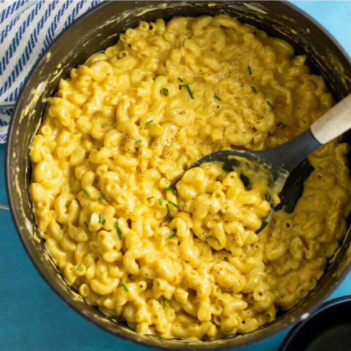 Large pot of stovetop mac and cheese made without flour with a wooden spoon in the middle on a bright blue background topped with fresh chives and black pepper
