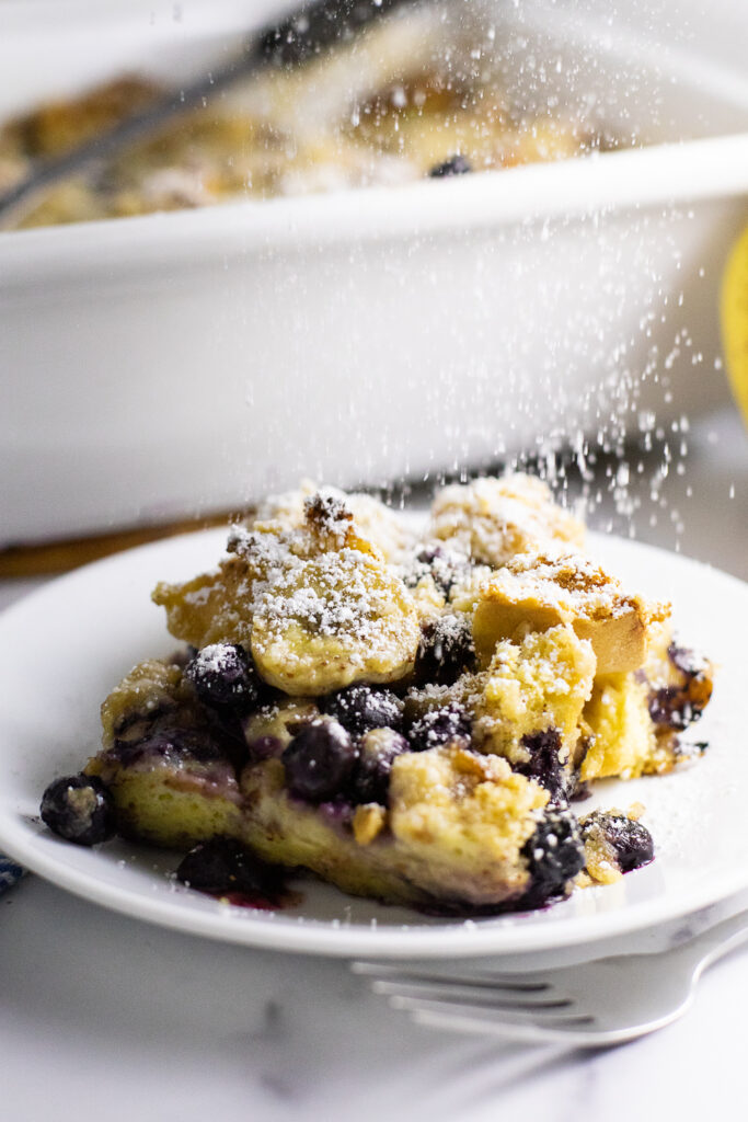 A piece of blueberry banana French toast bake on a small plate with powdered sugar being sprinkled on. Plate is in front of a large casserole dish and next to a fork