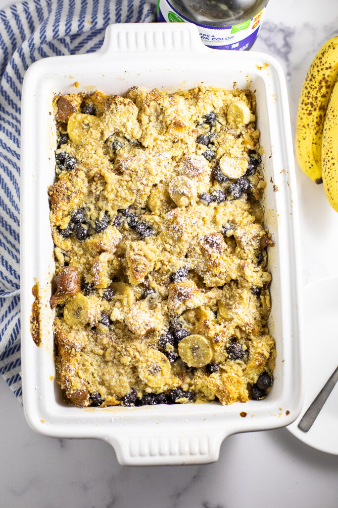 Blueberry Banana French Toast bake in a large white casserole dish topped with powdered sugar surrounded by a cloth napkin, bunch of bananas, and a bottle of maple syrup