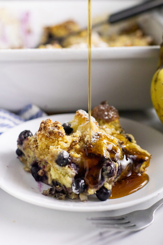 A serving of blueberry banana French toast bake on a small plate with syrup being drizzled on. Plate is in front a large casserole dish filled with the remaining dish.