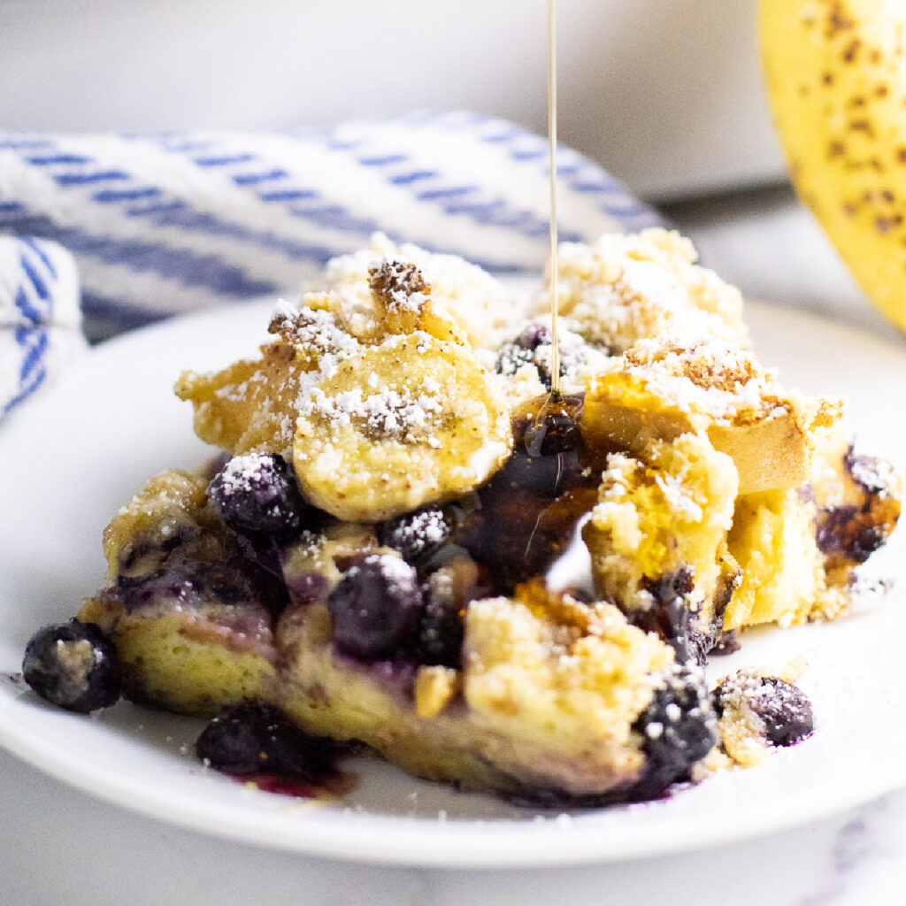 A piece of blueberry banana French toast bake on a small plate with syrup drizzling down on top