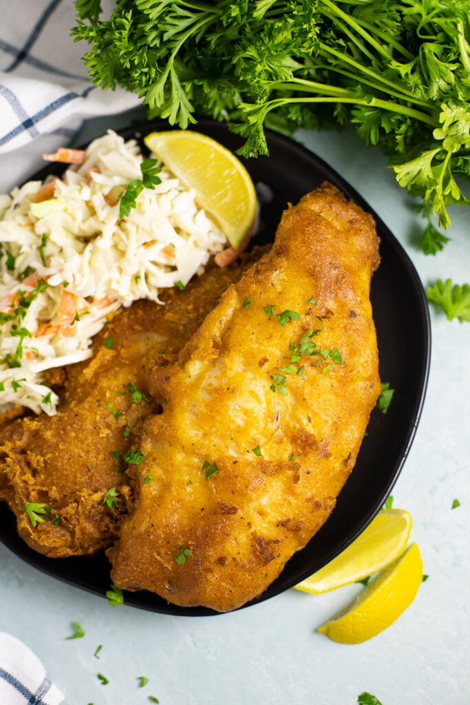 Two pieces of beer battered catfish on a plate with creamy coleslaw and a lemon wedge with fresh parsley and lemon wedges in the background around it.