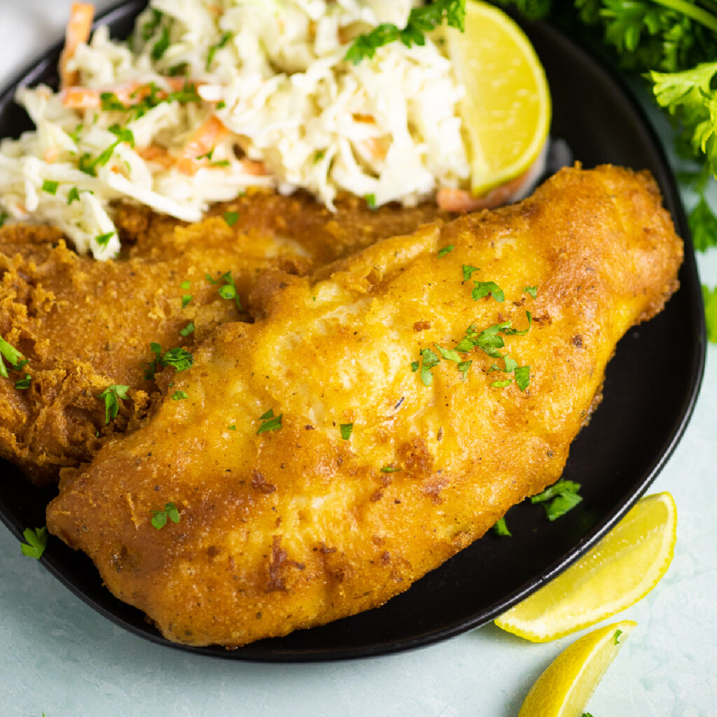 Two pieces of beer battered catfish on a plate with creamy coleslaw and a lemon wedge with fresh parsley and lemon wedges in the background around it.
