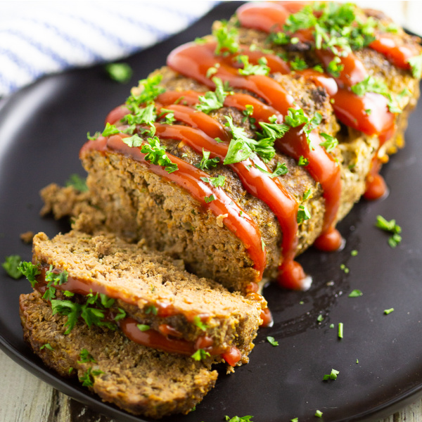 Full meatloaf on a large plate with 2 slices cut off garnished with ketchup drizzle and fresh chopped parsley.
