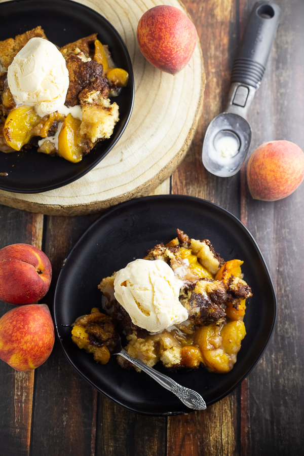 Overhead picture of peach cobbler topped with vanilla ice cream on a plate next to a spoon with another plate of peach cobbler next to it along with an ice cream scoop and 4 fresh peaches scattered around