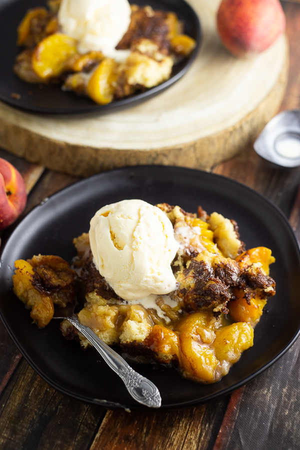 Peach cobbler on a plate with a spoon topped with ice cream with another plate of cobbler in the background with fresh peaches
