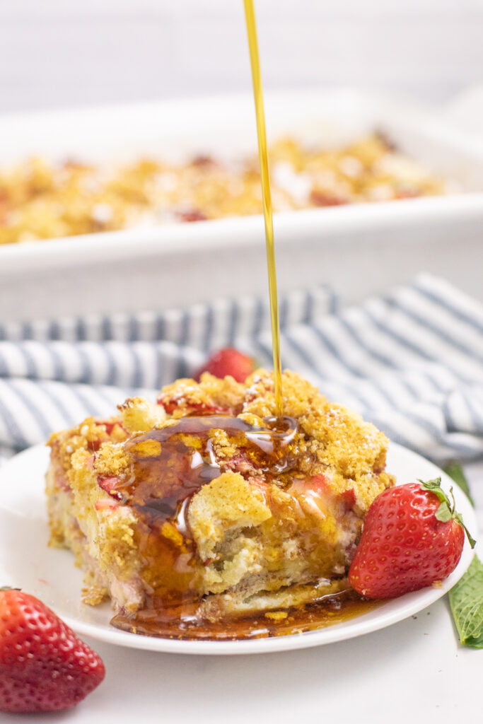 A piece of strawberry cream cheese French toast casserole on a small white plate next to a fresh strawberry with maple syrup drizzling on top and a linen napkin and the remaining casserole in the casserole dish behind.