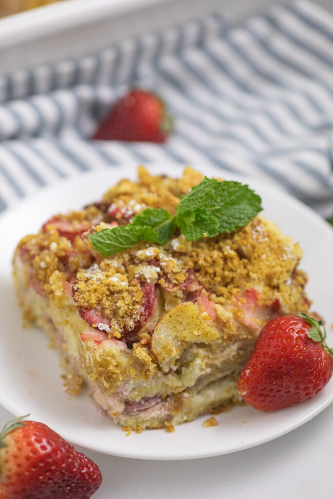 Strawberry cream cheese French toast casserole on a small plate topped with a mint sprig next to a fresh strawberry with more fresh strawberries and a linen napkin surrounding it.