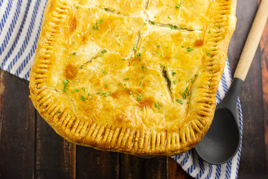 Fully baked golden chicken pot pie on a dark rustic wood background with a wooden spoon and striped cloth napkin.