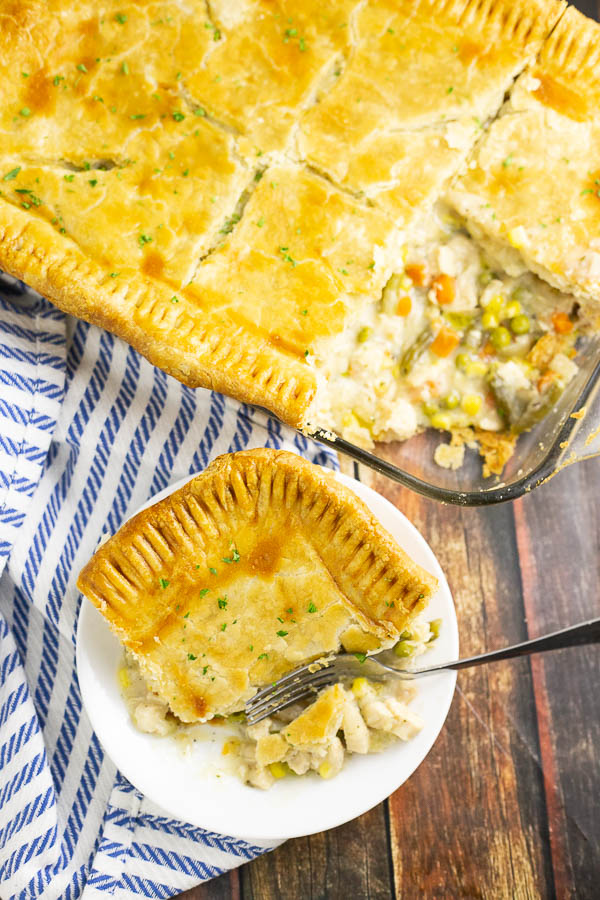 Corner slice of chicken pot pie on a white plate with a blue fork. Full pot pie is in the background sitting on a white and blue striped napkin