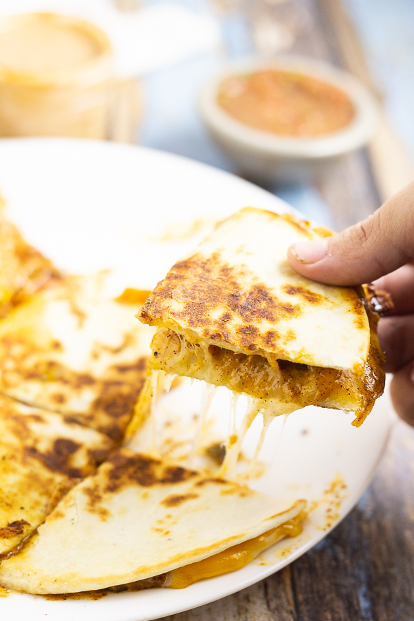 Hand picking up a quarter of a gooey, cheesy quesadilla from a white plate.