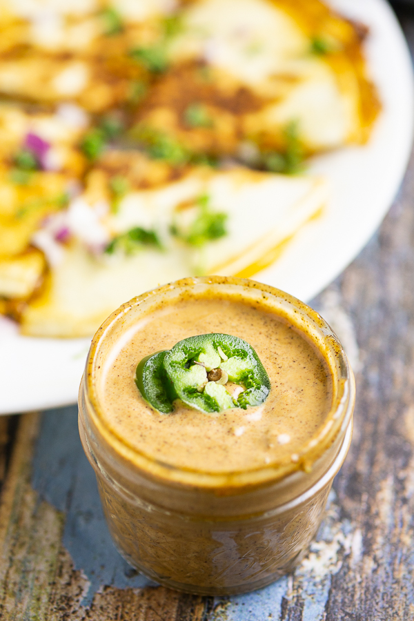 Copycat Taco Bell Quesadilla Sauce in a small glass jar topped with 2 jalapeno slices with a quesadilla in the background.