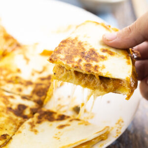 Hand picking up a quarter of a gooey, cheesy quesadilla from a white plate.