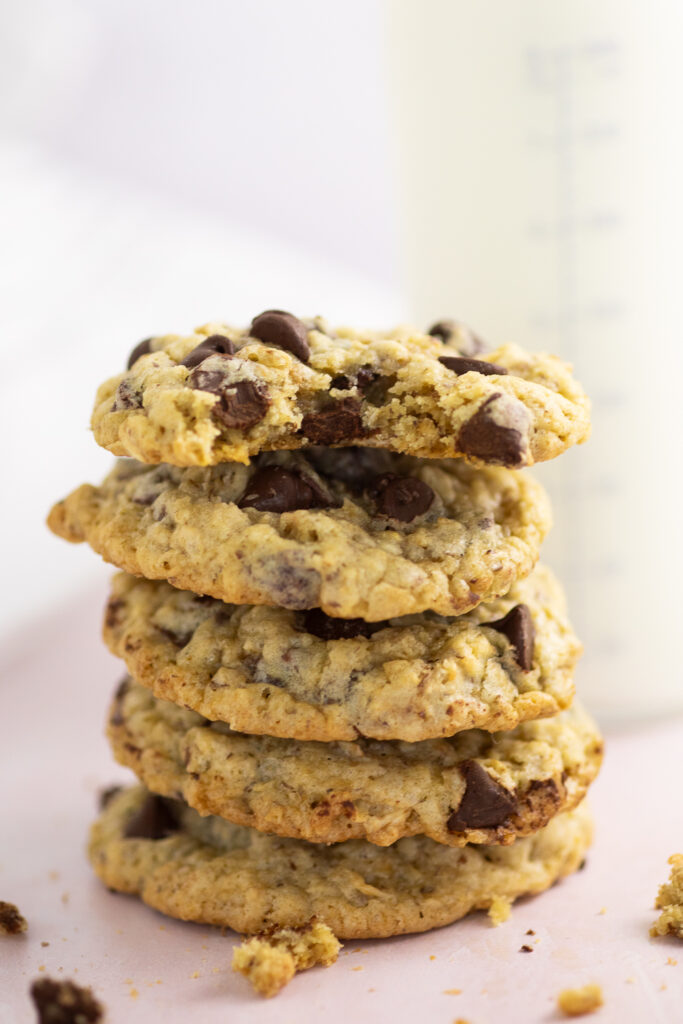 A stack of lactation cookies on a pink marble background with a bottle full of milk behind. The top cookie has a bite taken out.