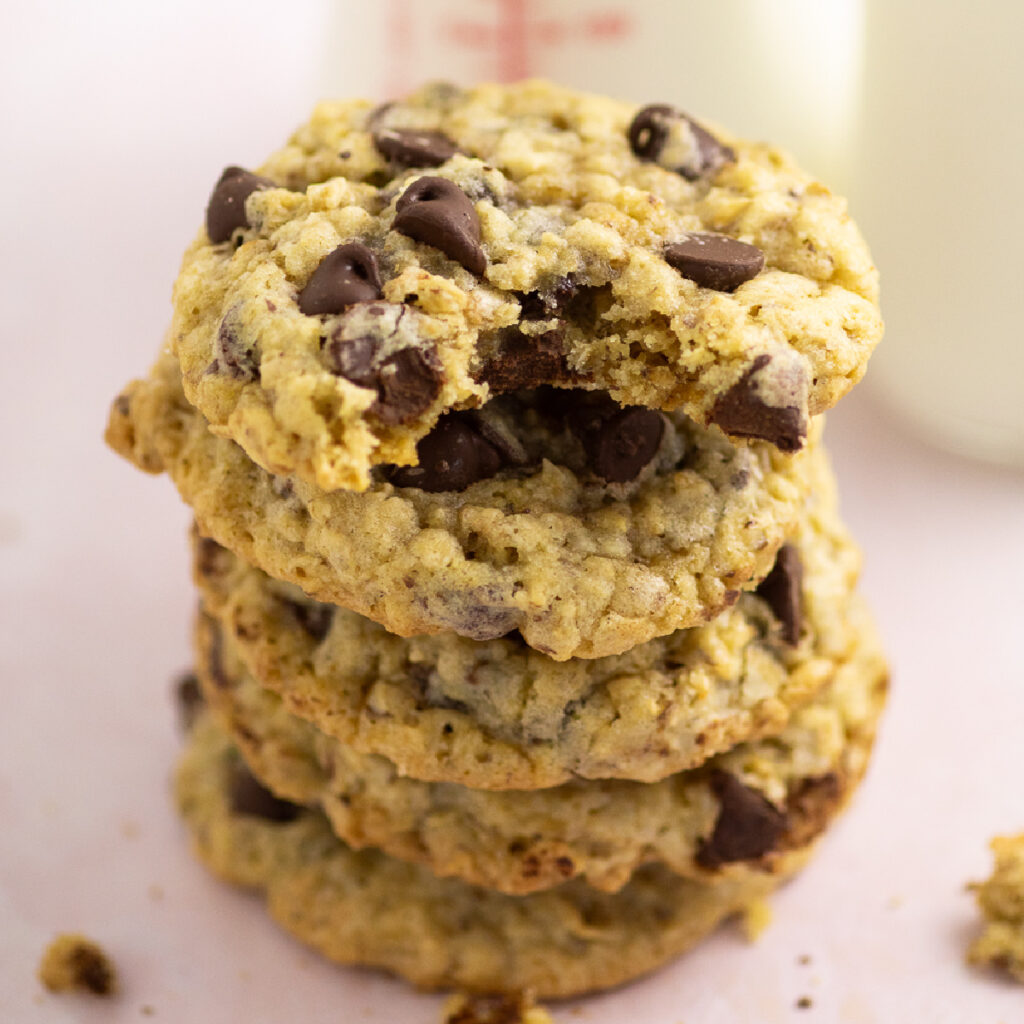 A stack of lactation cookies on a pink marble background with a bottle full of milk behind. The top cookie has a bite taken out.