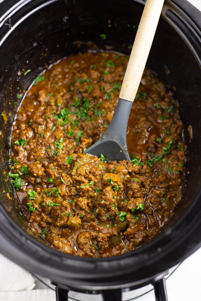 Sloppy joe filling topped with fresh chopped parsley with a wooden spoon sticking out in a large slow cooker