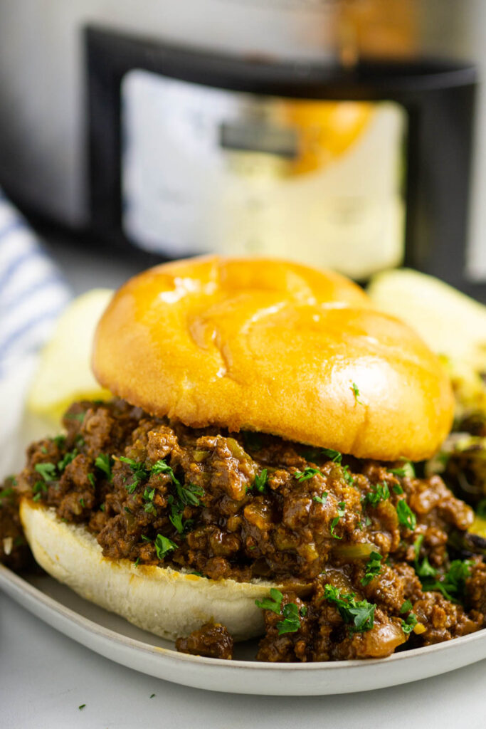 Sloppy joe with fresh chopped parsley and a toasted bun sitting on a small white plate in front of a Crock Pot