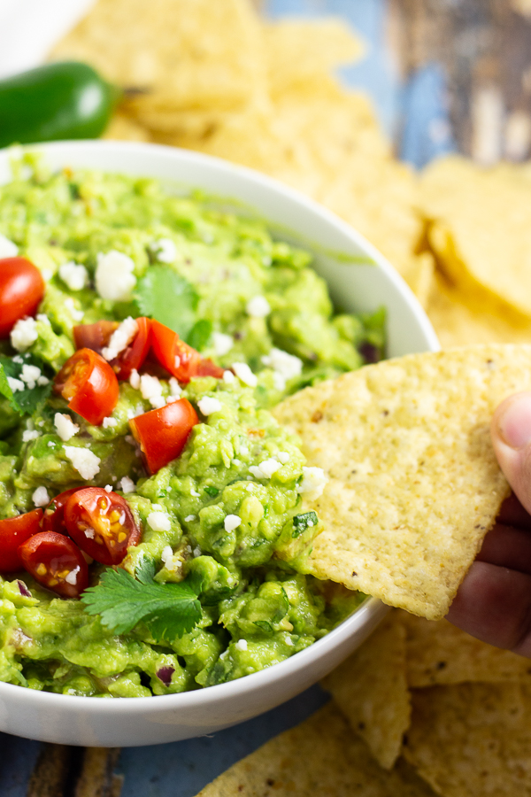 Guacamole in a bowl topped with halved cherry tomatoes, fresh cilantro, and cotija cheese with a chip dipping into and tortilla chips and a jalapeno in the background