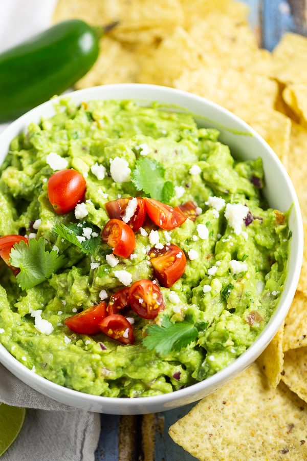 Guacamole in a bowl topped with halved cherry tomatoes, fresh cilantro, and cotija cheese with tortilla chips and a jalapeno in the background