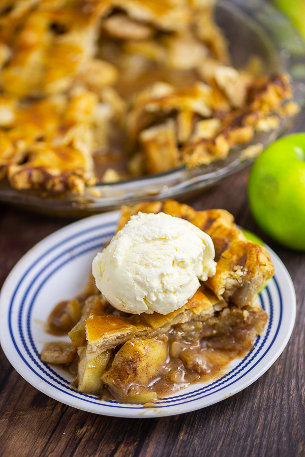 Slice of apple pie on a small plate topped with a scoop of vanilla ice cream. Green apple and pie in a pie dish are behind.