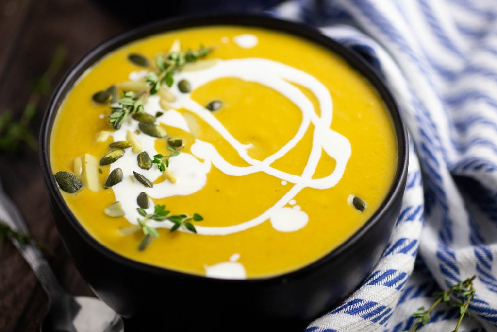 Butternut Squash Bisque in a matte black bowl with a swirl of cream and pepitas and fresh thyme on top sitting on a white and blue linen and a rustic wood background.