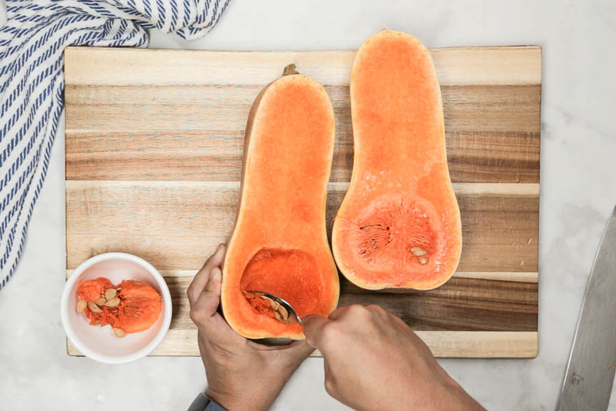 Large butternut squash cut in half on a cutting board with the pulp and seeds being scooped out.