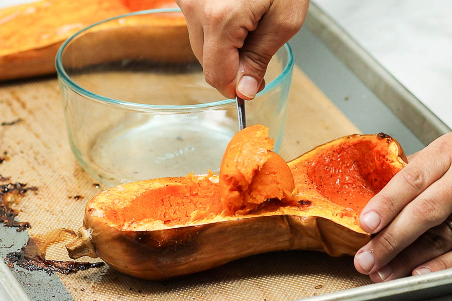 Roasted butternut squash on a baking sheet with the flesh being scooped out with a spoon into a glass bowl.