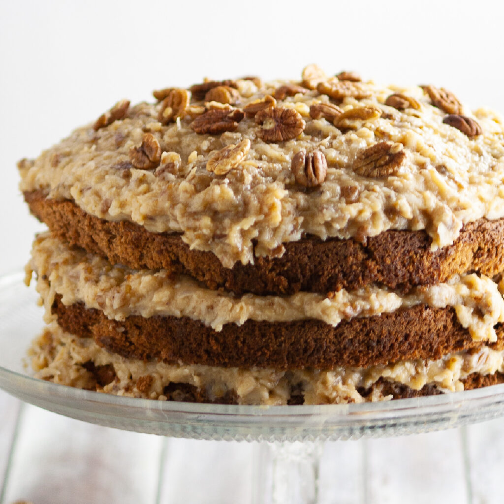 Three layer German Chocolate Cake topped with toasted pecans on a glass cake stand on a rustic wood background