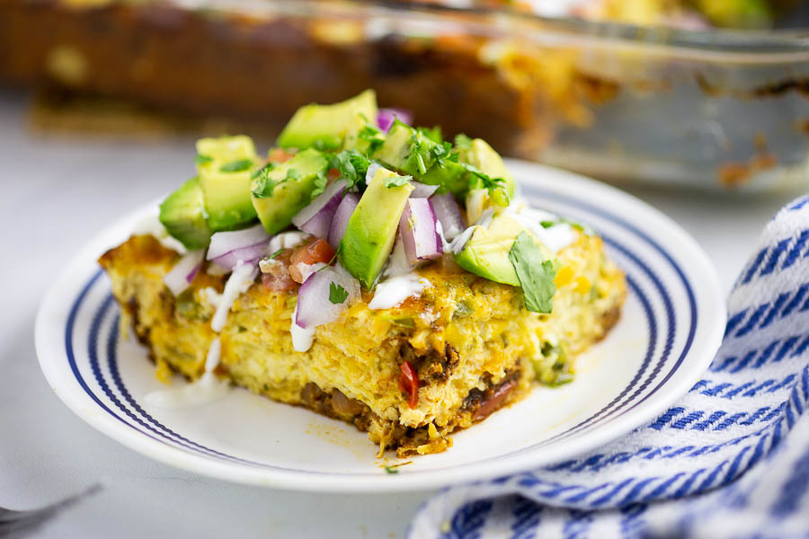 Mexican breakfast casserole on a small white and blue plate, topped with avocado next to blue and white linen and full casserole in the background.