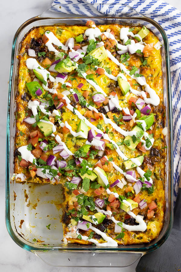 Mexican Breakfast Casserole topped with pico de gallo, red onion, avocado, and a drizzle of sour cream in a glass baking dish on a white marble background with a blue and white linen