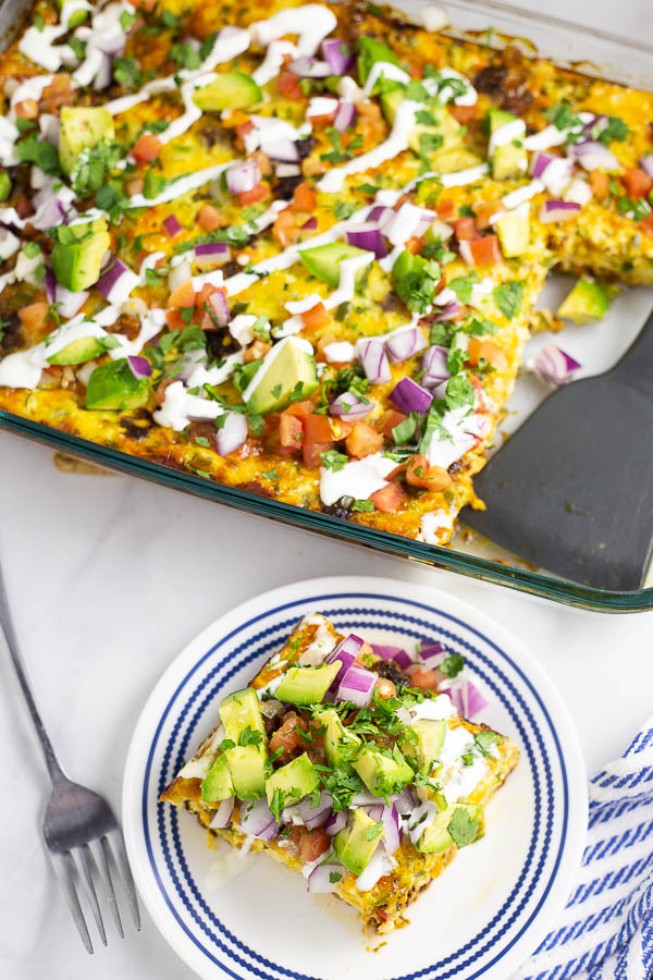 Overhead view of a piece of Mexican Breakfast Casserole on a small white plate topped with avocado, the full casserole with a gray spatula sitting in the glass baking dish sitting on a marble counter with blue and white linen.