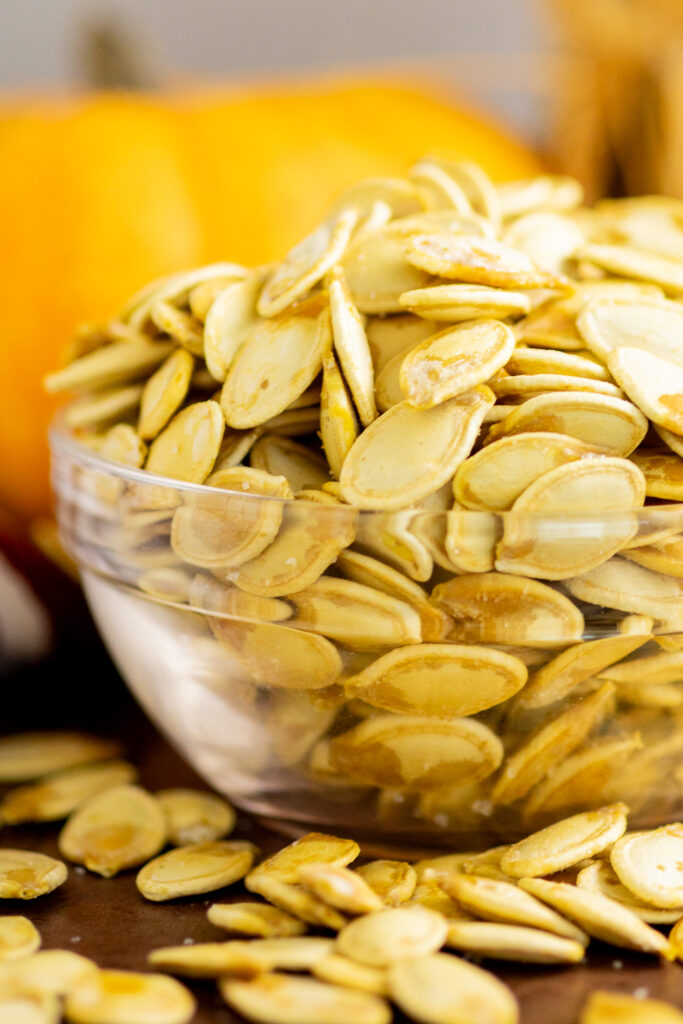 Close up of toasted pumpkin seeds in a glass bowl with more pumpkin seeds around it on a rustic wood background and a mini pumpkin behind.