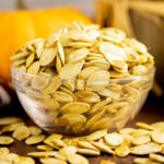 A small glass bowl full of toasted pumpkin seeds with more pumpkin seeds around it on a rustic wood background and a mini pumpkin behind.