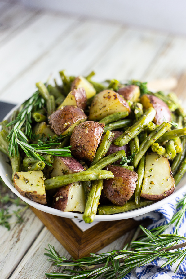 Roasted red potatoes and green beans in a white bowl with fresh rosemary sprigs on a white rustic wood background