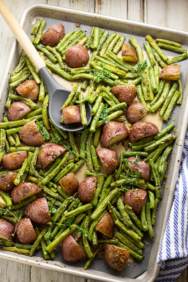 Roasted red potatoes and green beans on a large baking sheet with a gray silicone and wooden spoon on top on a white rustic wood background