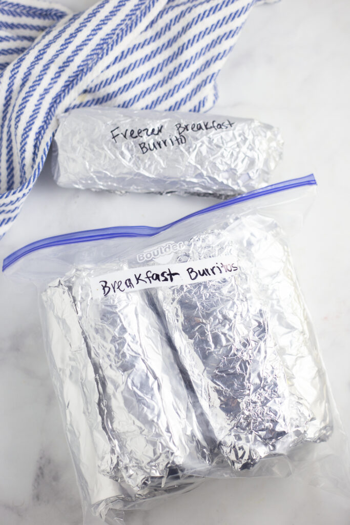 Freezer burritos wrapped in foil in a plastic zip baggy labeled "breakfast burritos" with one wrapped burrito not in the bag next to a kitchen towel on a white marble surface.