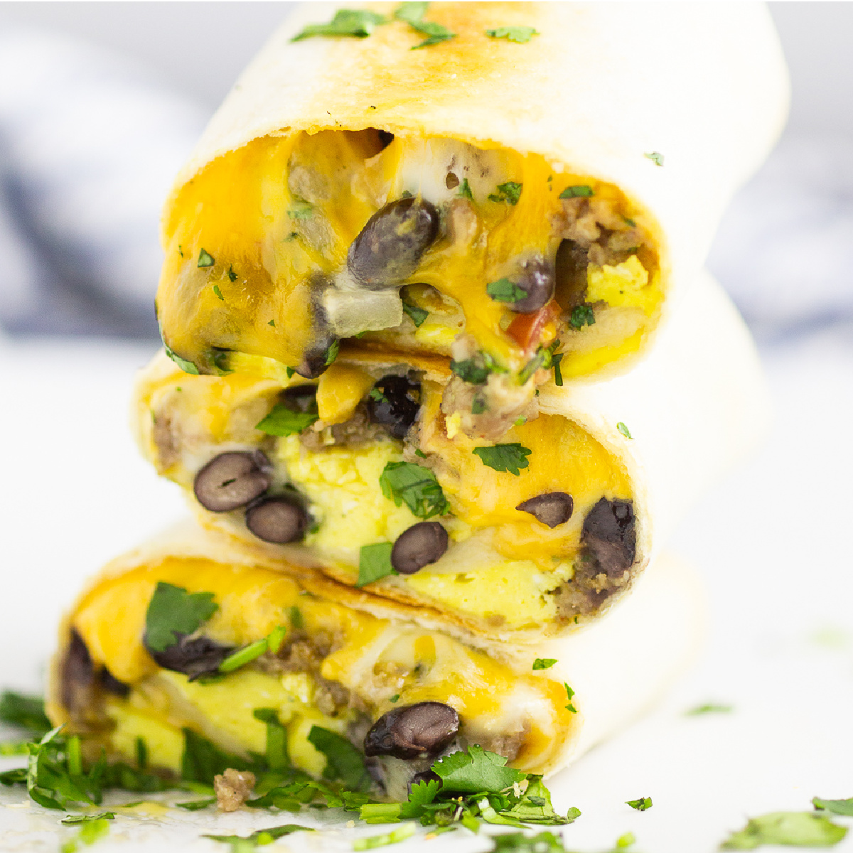 Stack of three halves of frozen breakfast burritos with the ingredients, including eggs, black beans, and cheese with cilantro sprinkled around on a white marble surface