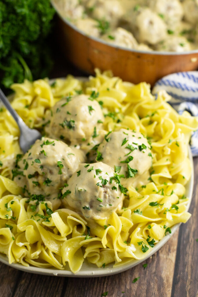Authentic Swedish meatballs on top of buttered egg noodles with creamy brown gravy and fresh parsley on top on a rustic wood background with a bunch of parsley behind.