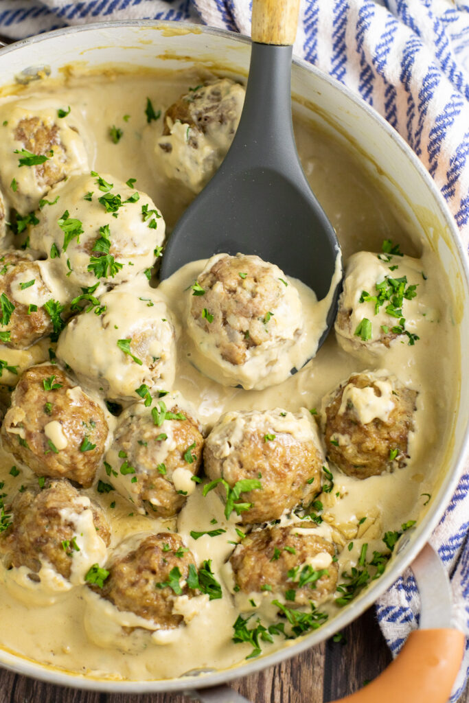 Authentic Swedish meatballs in a deep copper skillet smothered in creamy brown gravy and topped with fresh parsley with a spoon scooping up one meatball. The skillet is next to a blue and white striped linen napkin.