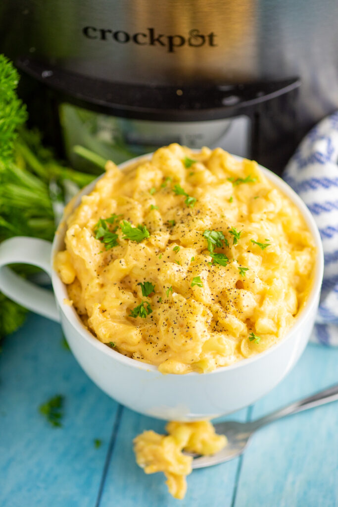 Overhead view of a big bowl full of slow cooker mac and cheese next to a fork with mac and cheese on it and in front of a Crock Pot and bunch of fresh parsley
