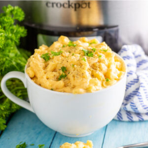 Macaroni and cheese in a big, deep bowl with a handle sitting in front of a Crock Pot, linen, and fresh bunch of parsley