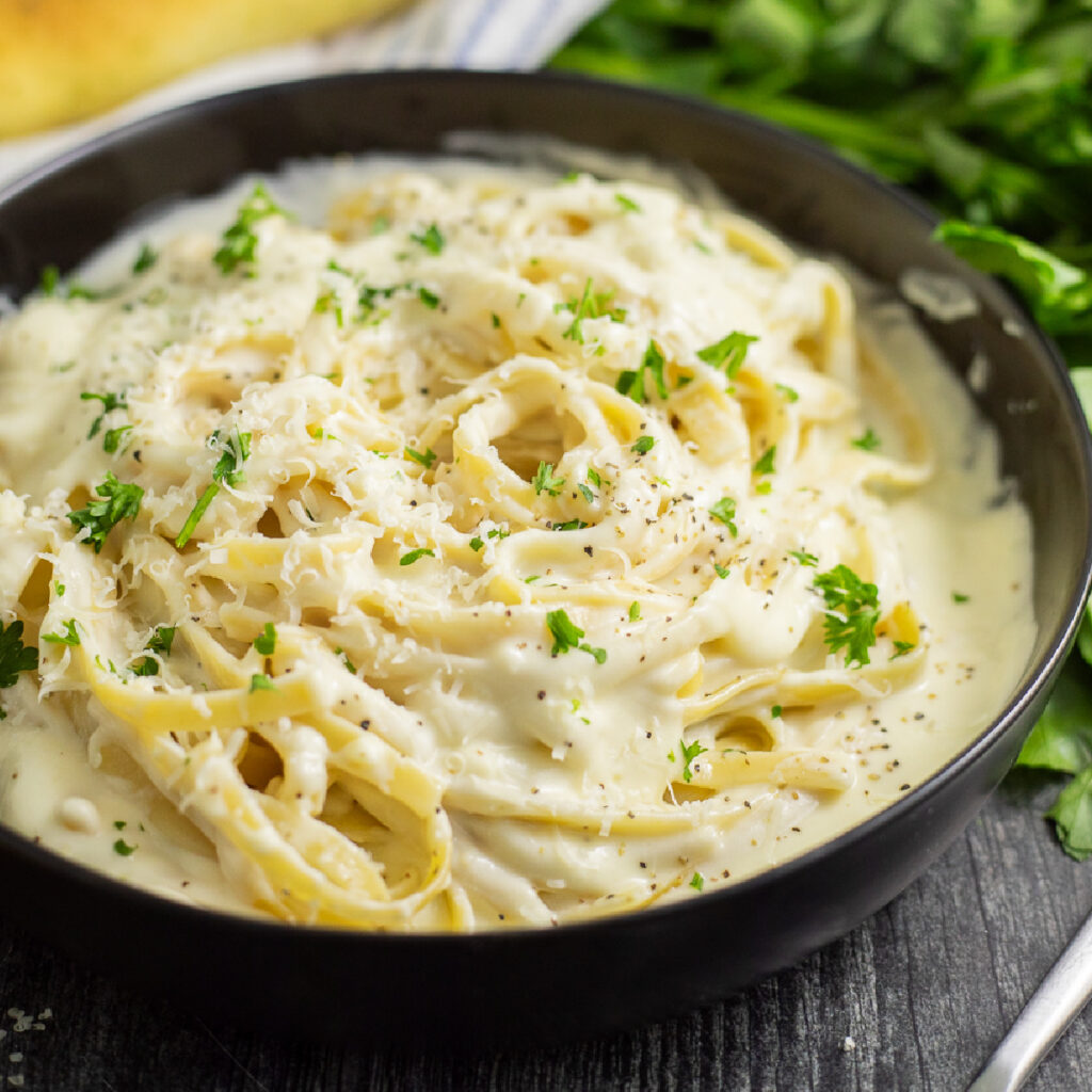 A black matte bowl of fettuccine alfredo topped with freshly chopped parsley on a rustic wood background with a bunch of fresh parsley, parmesan wedge, and a linen napkin behind.