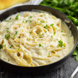 A black matte bowl of fettuccine alfredo topped with freshly chopped parsley on a rustic wood background with a bunch of fresh parsley, parmesan wedge, and a linen napkin behind.