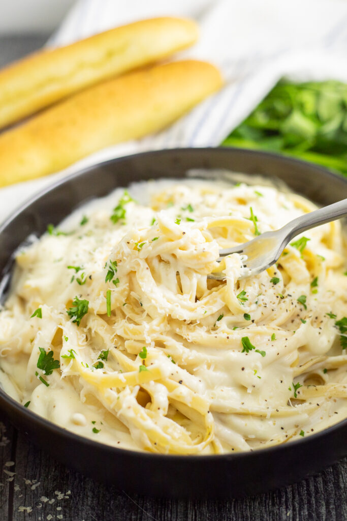 A black matte bowl of fettuccine alfredo topped with freshly chopped parsley on a rustic wood background with a bunch of fresh parsley, parmesan wedge, and a linen napkin behind.