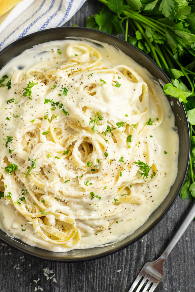 Overhead view of fettuccine alfredo topped with fresh parsley on a rustic wood background, surrounded by a fork, a bunch of fresh parsley, and a linen napkin.