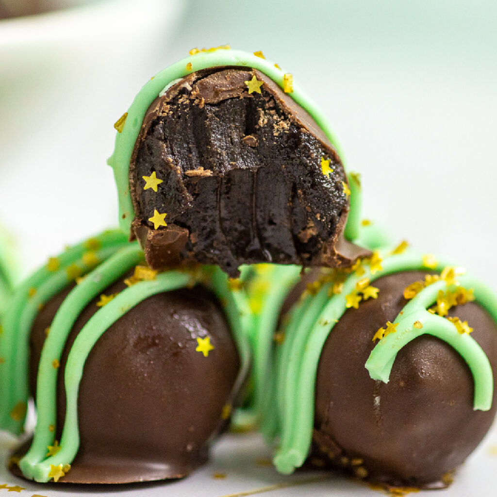 A stack of mint Oreo truffles with two on bottom and one on top. The one on top has a bite taken out of it. They're topped with green white chocolate and gold star sprinkles.