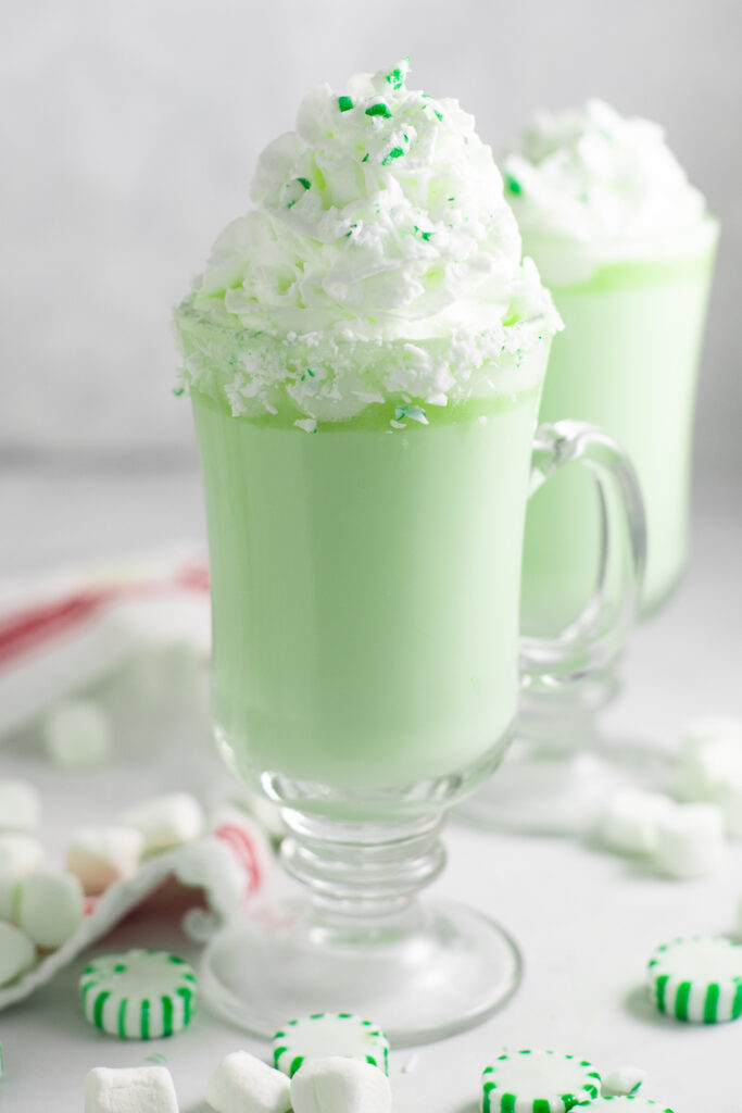 A glass mug of green mint white hot chocolate topped with whipped cream and crushed green mint candy pieces on a white marble surface surrounded by more star mint candies.