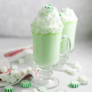 A glass mug of green mint white hot chocolate topped with whipped cream and crushed green mint candy pieces on a white marble surface surrounded by more star mint candies.